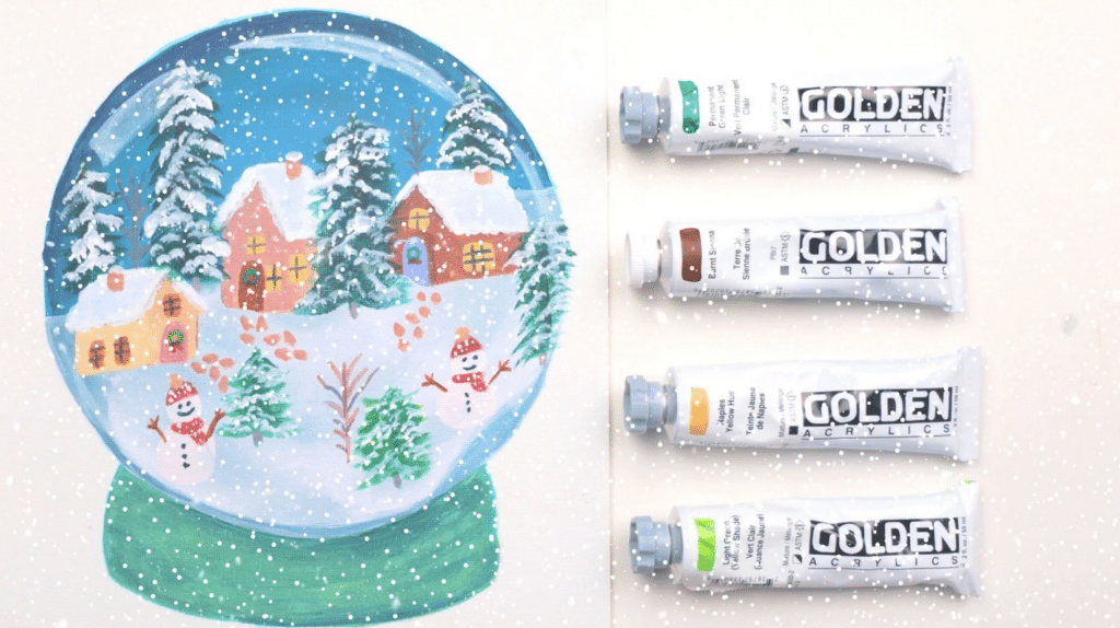 Four small tubes of acrylic paint laying next to a square white greeting card. The card is decorated with a painting of a snow globe containing snowmen, snowy evergreen trees and quaint houses.