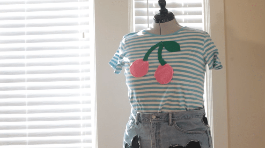 A mannequin wears a blue and white striped shirt with big appliqué cherries on the front and jeans. The mannequin sits in a room in front of two windows. 