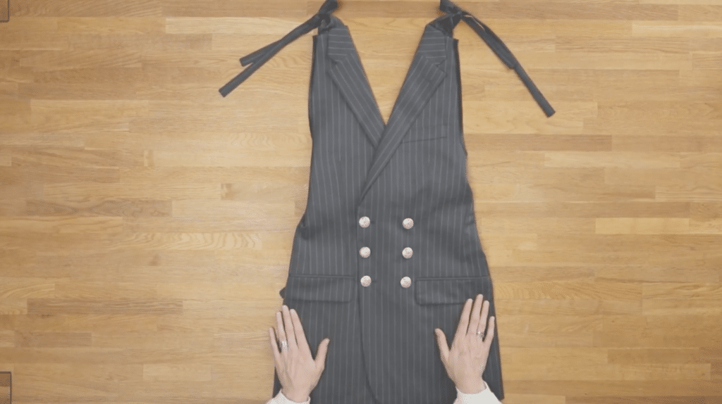 A dress lying on a wooden floor. The dress has six silver buttons near the waist in two vertical lines of three. The dress is sleeveless and short and made out of a dark gray striped suit jacket. Two hands are touching the dress ,palms down near the bottom hem. 