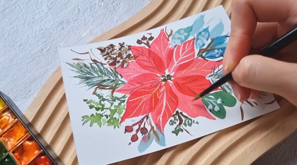 A person’s hand using a paintbrush to put the finishing touches on a watercolor holiday card. The card’s design depicts a red poinsettia flower surrounded by brown pinecones, pine sprigs, red berries and green leaves.