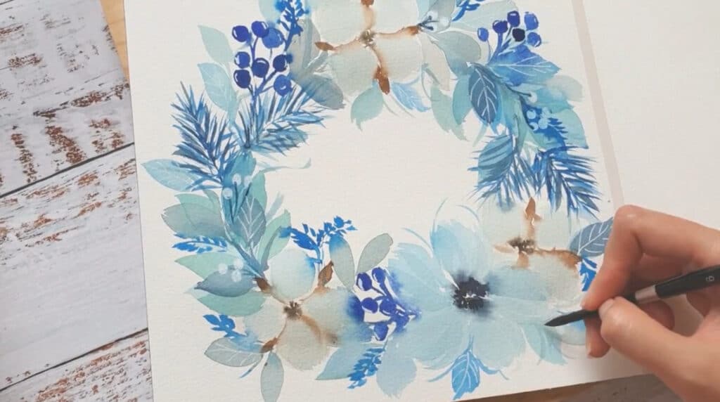 A person’s hand using a paintbrush to paint a watercolor image of a blue-toned wreath on a white greeting card.