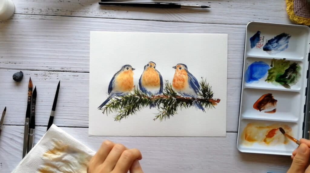 A rectangular white Christmas card laying on a white painted tabletop. The card features a watercolor illustration of three robins perched on a pine branch. A person’s hand is using a paintbrush to dip into a watercolor palette to the right of the card.