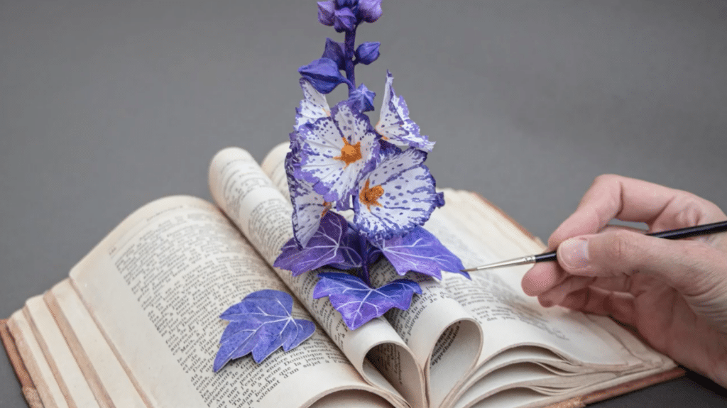 A stem made of paper and air dry clay and covered in flowers sprouts from an open book. The flowers are white and purple, and they grow from a purple stem covered in leaves and buds. A hand holding a black paint brush paints one of the purple leaves. 