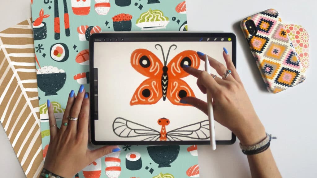 A woman’s hands holding an iPad that’s laying on a white tabletop. She’s using an Apple Pencil to complete a digital illustration of a butterfly, which has orange wings accented with black dots and white swirls.