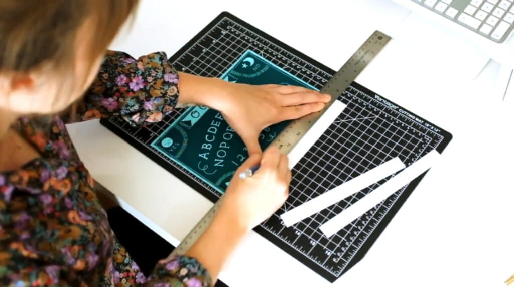 A woman’s hands using a metal ruler to create a straight line against a print of a Ouija board. With her right hand, she’s cutting the edge of the paper off with a utility knife. The print is laying on a black and white cutting mat, which is itself laying on a white tabletop.