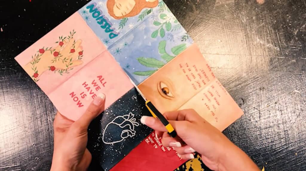 A woman’s hands using a pair of scissors to cut down the middle of a piece of paper. The paper is printed with eight different art pieces, four on one side and four on the other. Each piece of art is rectangular so that it can be cut into a page.