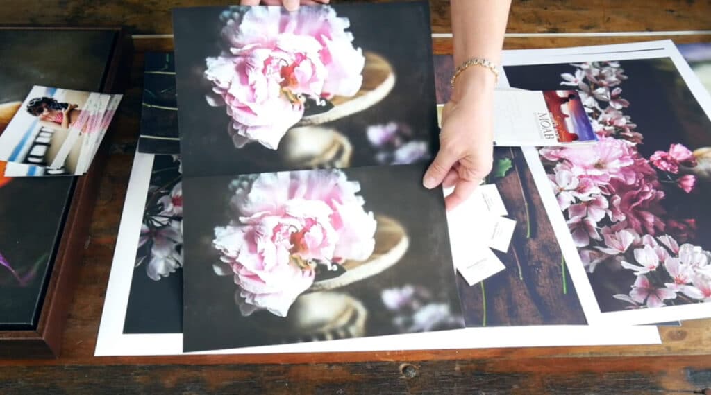 A woman’s hands holding two prints above a wooden tabletop. The prints are identical, and they both consist of an up-close photo of a pink rose. However, the print on the top is darker and more saturated, while the print on the bottom has a more muted and washed-out color palette.