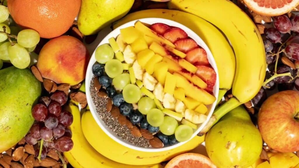 Overhead view of a pleasingly arranged bowl of sliced fruit, nuts and seeds surrounded by whole bananas, apples, grapes, citrus, peaches and nuts