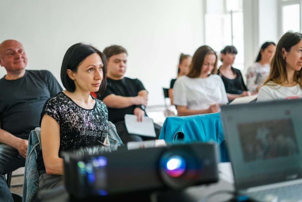 A group of eight people are sitting in a lecture-style setting. Some are looking up at a projected screen, while others are taking notes.