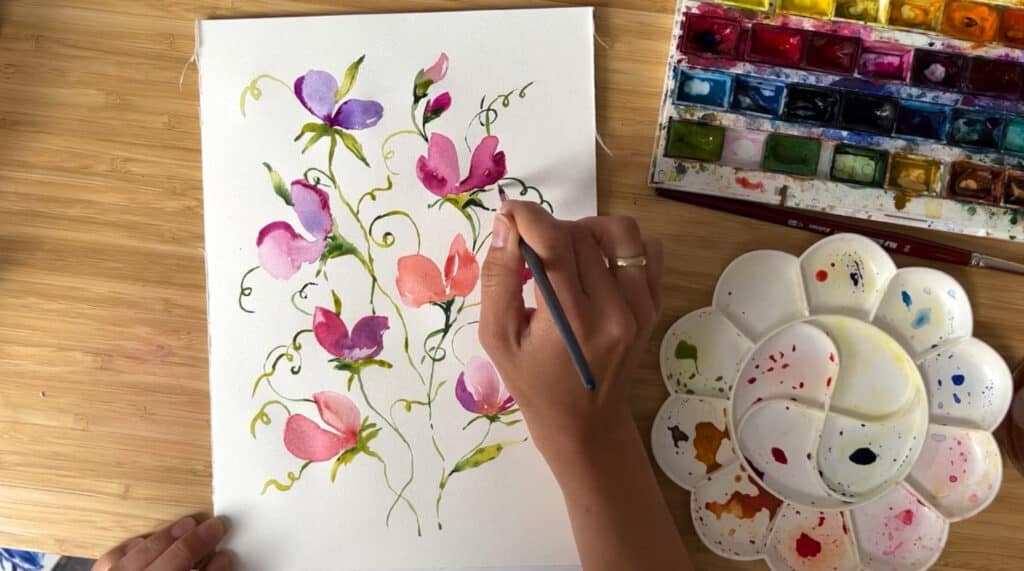 A person’s hand painting pink and purple flowers and green vines on a piece of paper. To the right of the painting in a round white watercolor palette and a box of watercolor paints.