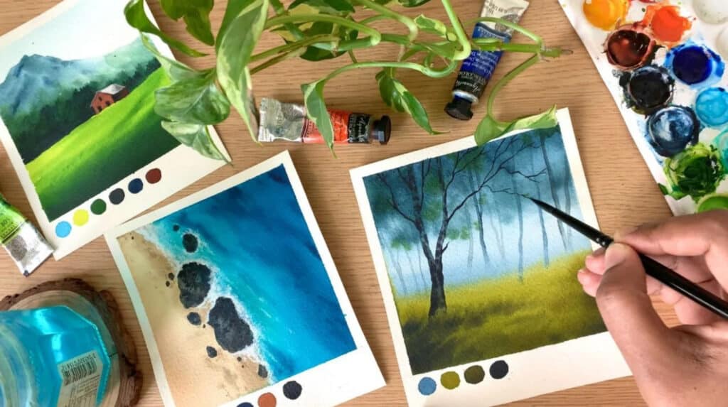 A person’s hand painting a small watercolor landscape painting of a tree in a grassy field. Also visible on the tabletop are two more paintings, one of a red barn in a green field, and another of an aerial view of a beach.