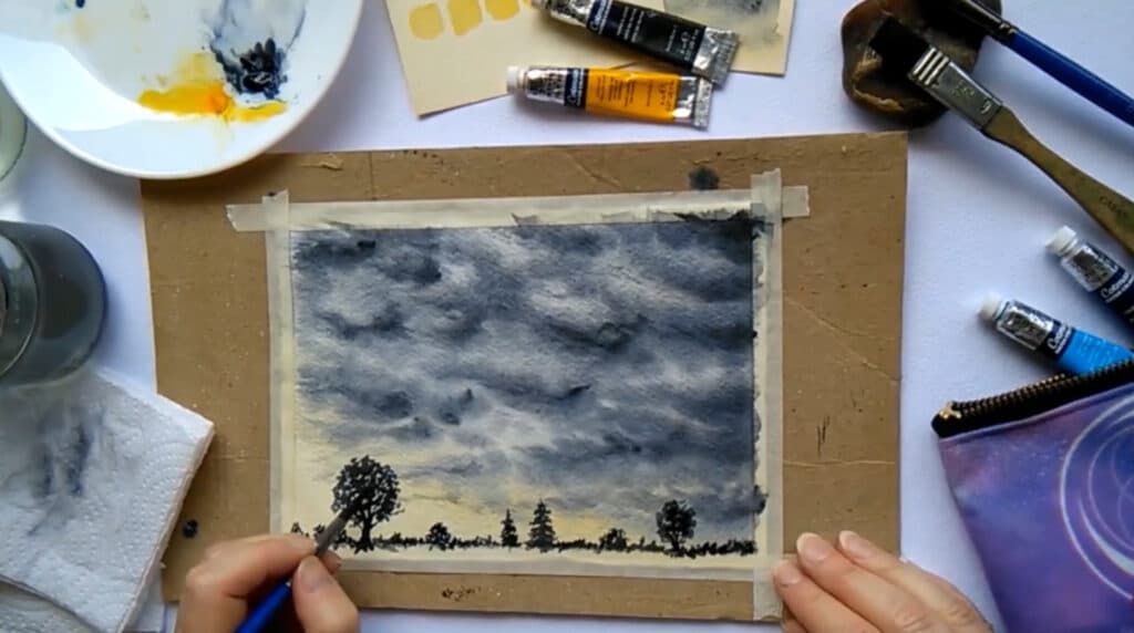 A person’s hand using a paintbrush to complete a watercolor painting of dark storm clouds over silhouetted trees. Also visible on the white tabletop are tubes of paint, a mug of water, paintbrushes and a round white paint palette.