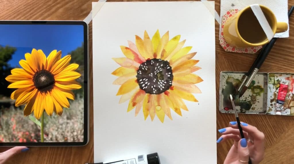 A person’s hand holding a paintbrush over a watercolor painting of a sunflower. To the left of the painting is an iPad displaying a reference photo, and to the right is a paint palette and mug of water.
