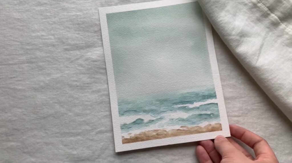 A person’s hand holding a small watercolor painting of a bluish-green sea underneath a muted blue sky.