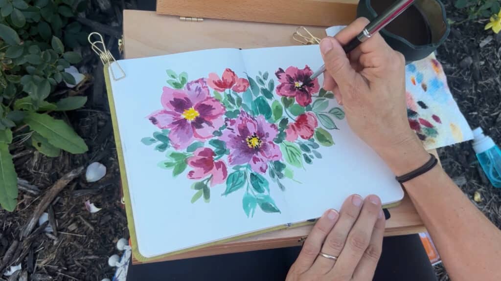 A person creating a watercolor painting in an outdoor garden. In a white sketchbook, they’re painting several red and purple flowers surrounded by green foliage.