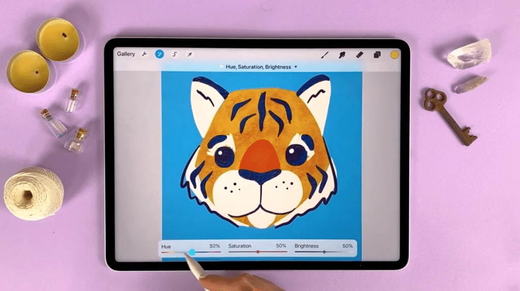 An iPad sits on a purple background surrounded by candles, thread and other miscellaneous objects. The iPad is open to Procreate. On the canvas is an illustrated tiger head on a blue background. A hand, holding a stylus, adjusts the hue by clicking on a sliding bar at the bottom of the screen.