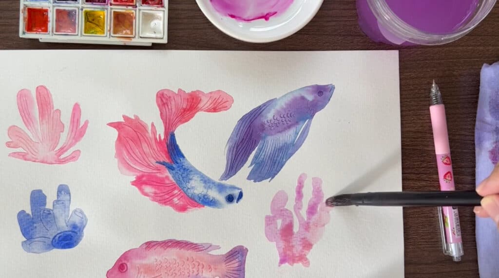A person’s hand using a paintbrush to paint a watercolor painting of several pink, blue and purple betta fish surrounded by clusters of matching coral.