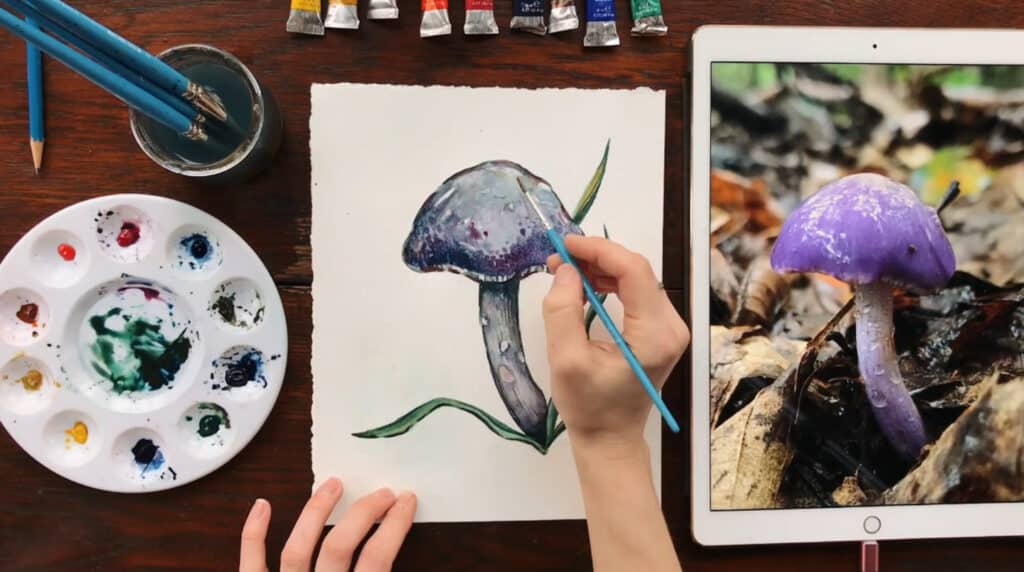 A person’s hand using a paintbrush to paint a purple mushroom on a white piece of watercolor paper. To the left of their painting is a round circular paint palette, and to the right is an iPad that’s displaying a reference photo of a purple mushroom.
