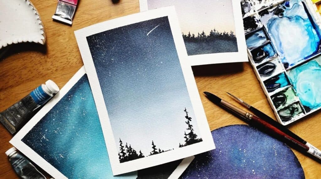 Several watercolor paintings of starry nights skies strewn on a wooden tabletop. They are surrounded by tubes of paint, paintbrushes and a white rectangular paint palette filled with various shades of blue paint.