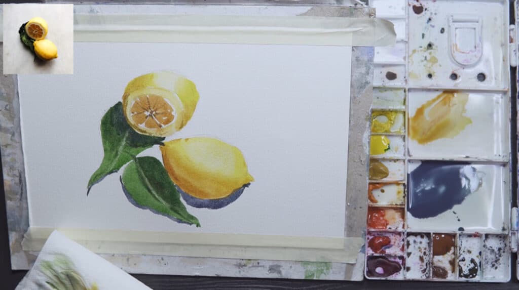 A watercolor painting of two yellow lemons with green leaves. To the right of the painting is a rectangular white paint palette. In the top-left corner, a reference photo is visible.