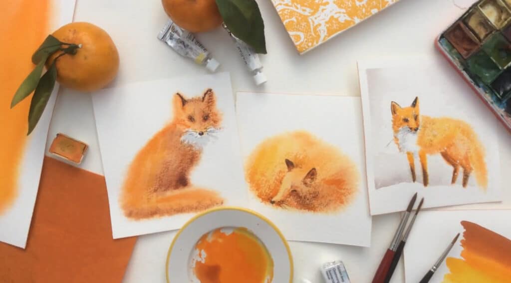 Several watercolor paintings of fluffy, bright orange foxes laying on a tabletop. They are surrounded by brushes and tubes of paint, as well as a couple tangerines.