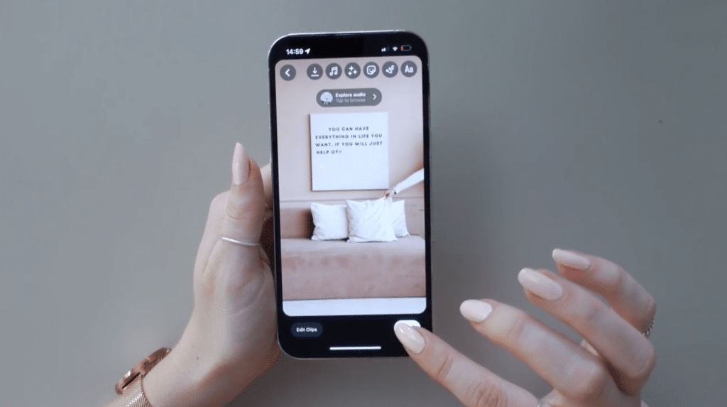 A phone is open to an Instagram reel that the creator is editing. One hand is holding the phone and the other is clicking a button on the screen. The video clip on the phone shows a hand placing a pillow onto a couch with a text-covered canvas hanging on the wall.