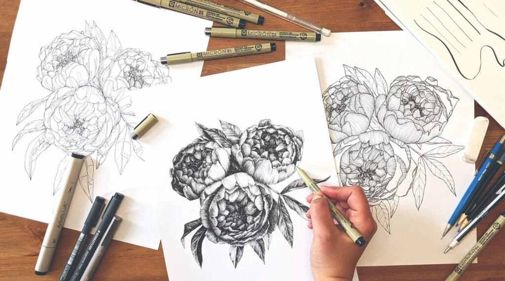 A person using ink pens to draw realistic flowers on a sheet of white paper. They’re doing so on a wooden tabletop that’s filled with pens, pencils and sketches of the flowers.