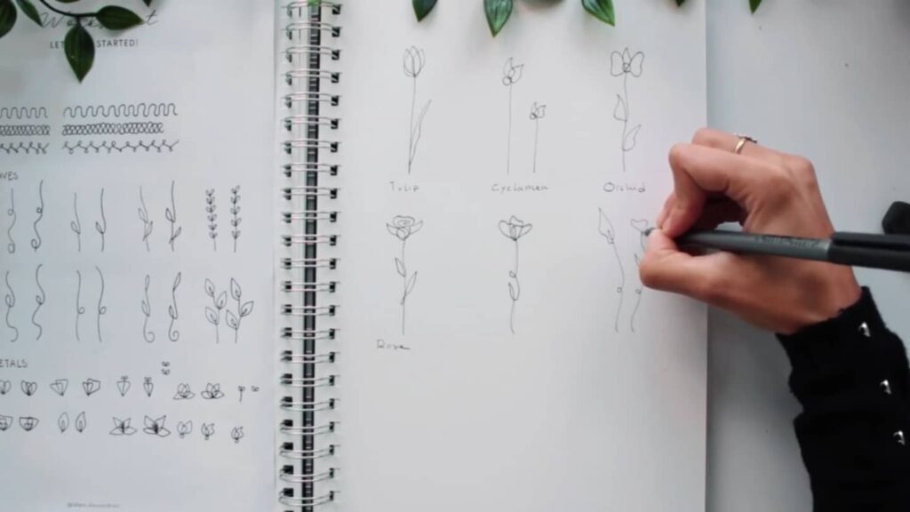 Open spiral sketchbook with artists’ hand drawing several designs and flowers in single, continuous lines.