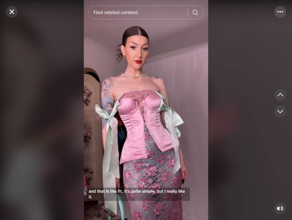 A woman standing inside a room with white walls. She’s wearing a pink silk corset over a pink lacy dress, and has decorated the top of the corset with large mint green silk bows. Behind her, a vintage-style folding screen decorated with bouquets of pink roses is visible. 