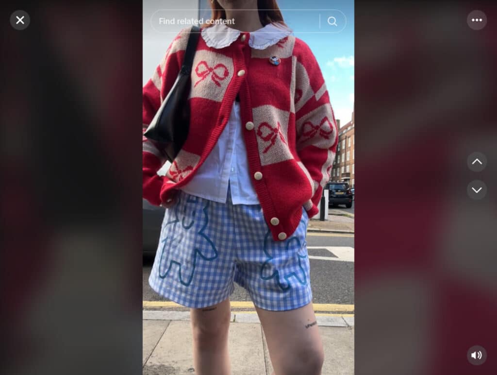 A woman standing on a city sidewalk with a street in the background. She’s wearing a red and beige square print cardigan that’s decorated with red bows, and white and blue gingham shorts that are decorated with a teddy bear motif. 