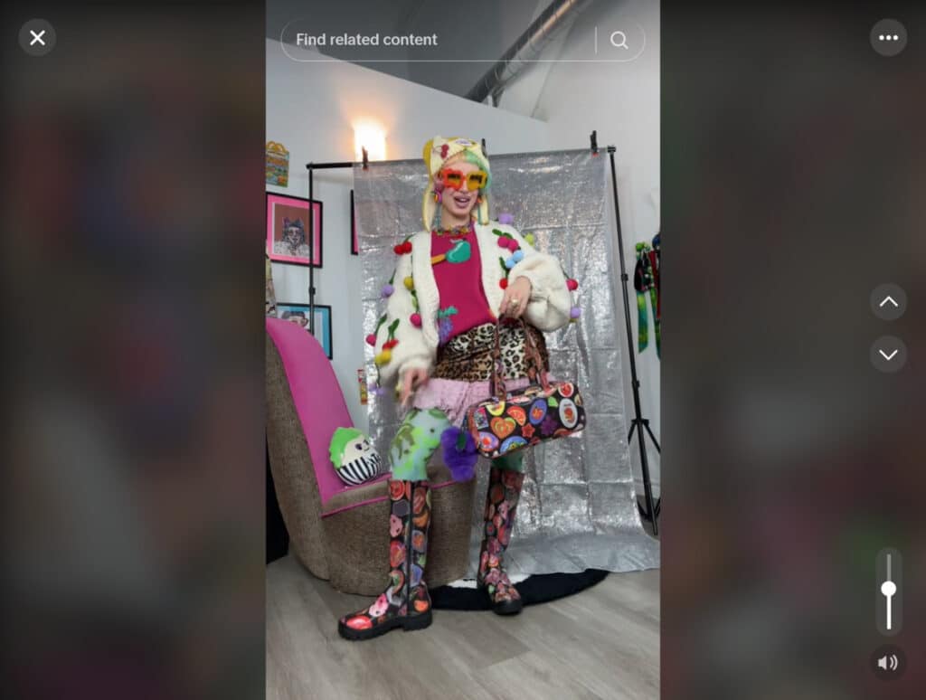 A woman standing in front of a silver photo backdrop in an apartment with white walls. She’s wearing a loud, colorful outfit that includes a fruit-themed cardigan, hat, purse, glasses, earrings and boots. 