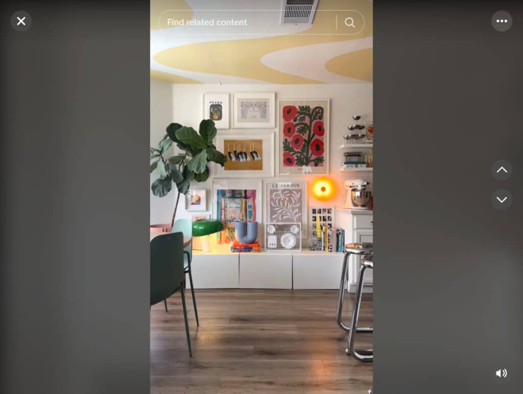 An apartment living room with wood floors and white walls. The visible wall is decorated with several framed art pieces, a warm amber-colored light, several books, sculptures and other pieces of decor, and a large potted plant. 