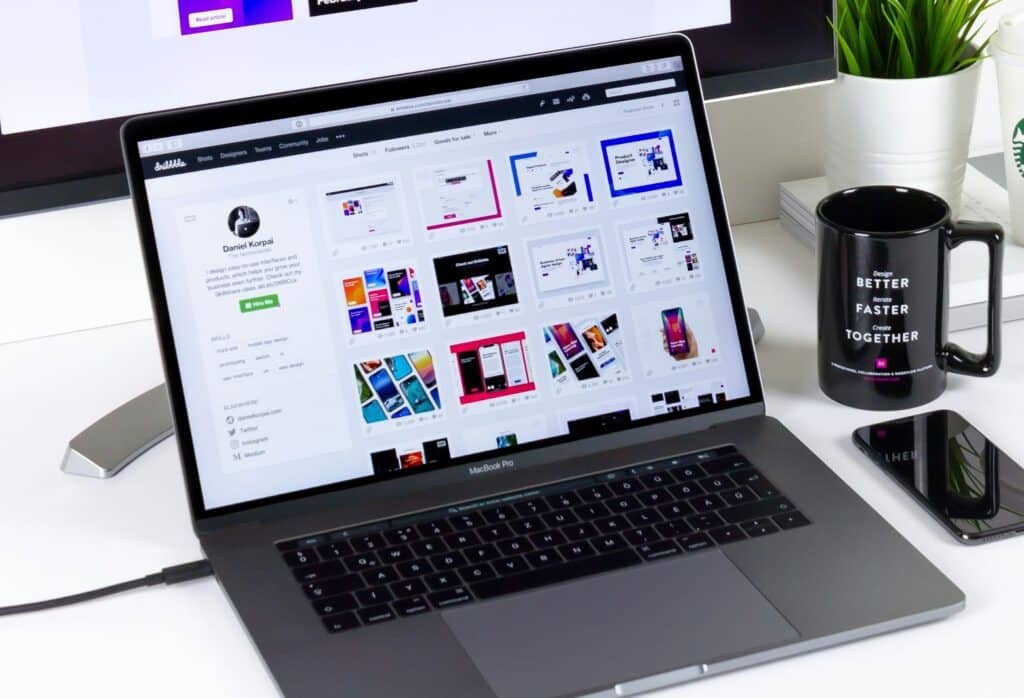 A laptop sits on a white table next to a phone and a black mug. The laptop is open to the portfolio site Dribbble. The page shows the portfolio of a designer named Daniel Korpai, which features his bio, his social media links and pictures of some of his most recent designs.