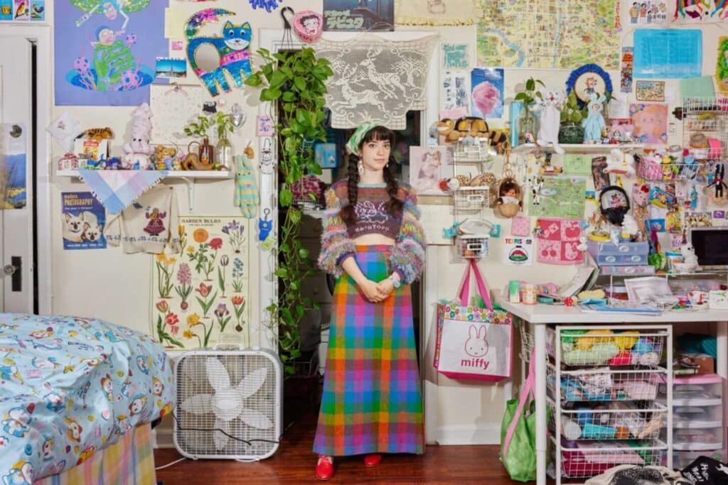 A woman dressed in a brightly multicolored outfit standing in a bedroom. The room’s walls are decorated with layers of posters, photos, hanging objects and shelves of trinkets. The desk is filled with colorful crafting materials, and the bed features cartoon-themed bedding. 