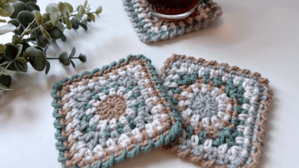 In the foreground, two crocheted "granny squares." A plant adds texture to the left, and a third granny square is mostly off-screen at the top of the image, being used as a coaster with a drink resting on it.
