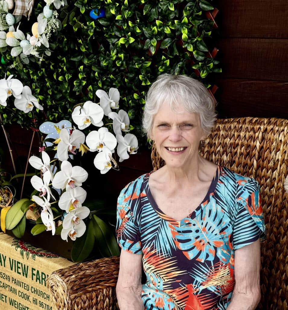 A woman sits in a chair and smiles at the camera. Behind her, white orchids bloom.