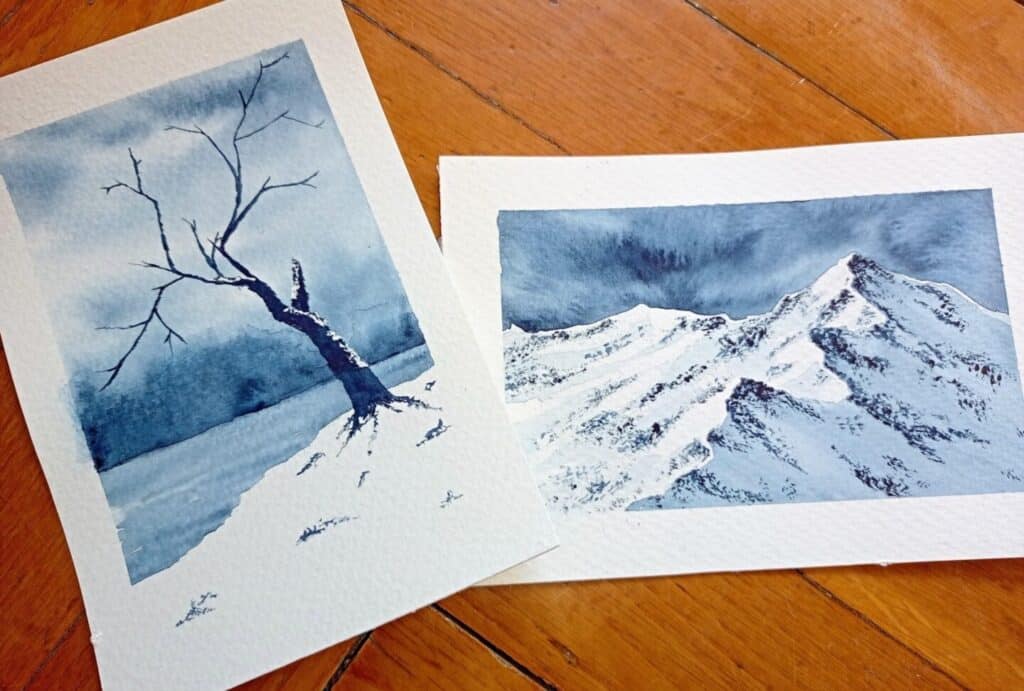 Two watercolor paintings laying next to each other on a wooden surface. One depicts a bare tree in the middle of a stark snowy landscape, and the other depicts snowy mountains under a cloudy sky. Both are painted in shades of blue.