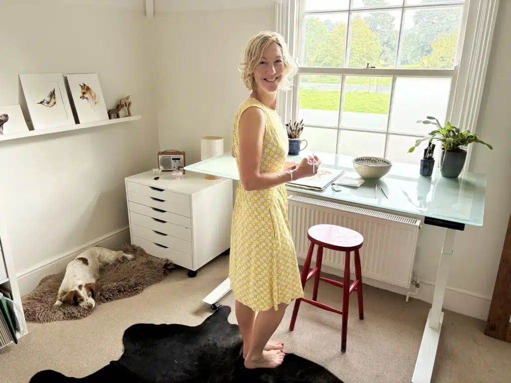 The watercolor artist Jane Davies standing in her painting studio. She’s wearing a yellow sundress and painting at a standing desk. Some of her watercolor paintings are hanging on the wall. Her pet Spaniel is sleeping on a fluffy bed on the floor.
