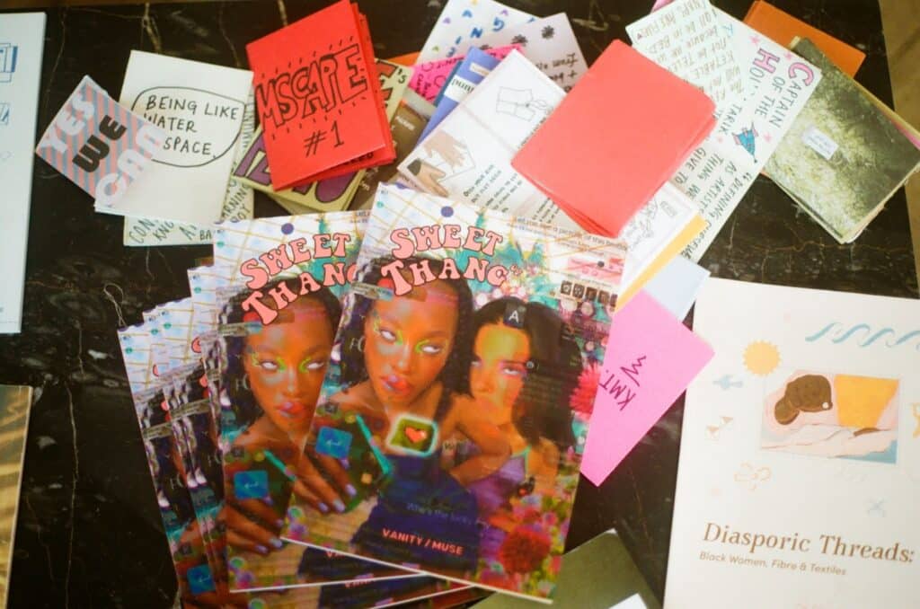 Multiple copies of a zine titled “Sweet Thang” and features two girls on the cover sits on a table surrounded by other zines.