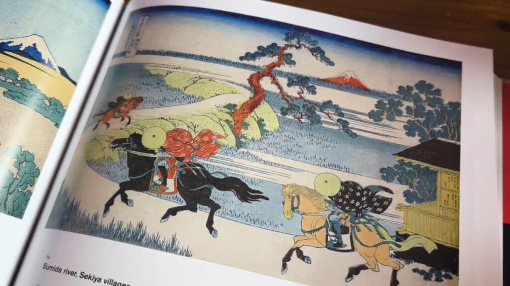 An art book open to a page displaying the print ‘Sumida River Sekiya Villages’ from Hokusai's ‘Thirty-six Views of Mount Fuji’ series. The print depicts people riding on horseback across a grassy field with Mount Fuji in the background.