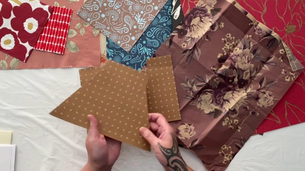 View of a worktable from above, an artist’s hands holding a piece of tan paper covered in a grid of small, white, oblong shapes, and the white table covered in layers of colorful decorative papers of red and purple florals, blue and brown paisleys and pinks with green leaves