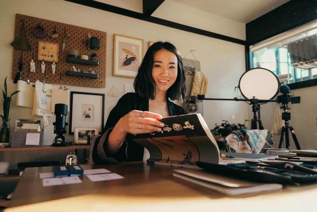 A woman sitting at a brown wooden desk in a room with white paint on the walls. She is turning the page of an art book. On the wall behind her is a pegboard filled with art supplies. The window on the right side of the room is letting in natural light.