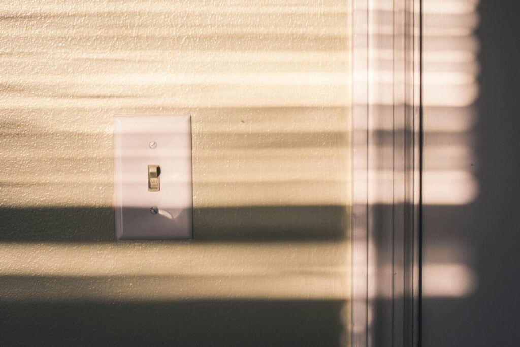 A photo of a white light switch on a beige painted wall. There is golden sunlight falling across the light switch in horizontal stripes, as if it’s streaming through window blinds.