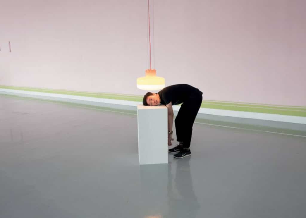 A man standing in a large, mostly empty art gallery room. He is bent over at the hips so his head is resting on a white rectangular art pedestal. Directly over ahead is a ceiling lamp that’s hanging about an inch from his face. 