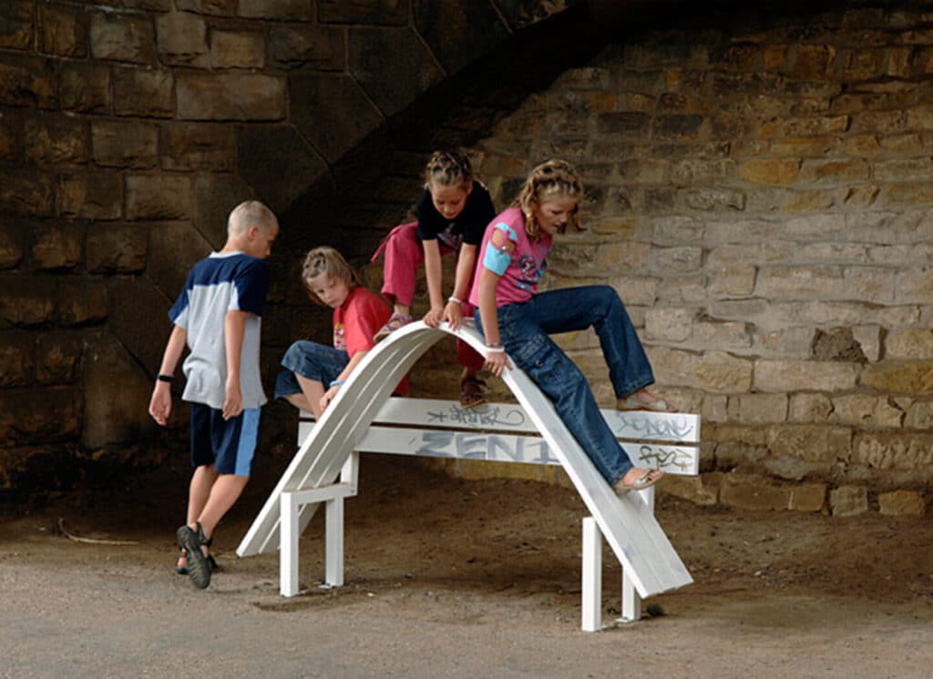 A group of children play on a bench that’s had its seat curved upwards so it forms an arch.