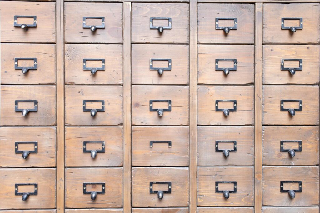 Twenty-five brown wooden drawers in a grid pattern. Each one is affixed with a metal library-style drawer pull. One of the drawer pulls is broken off.