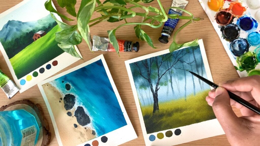 Flatlay photo of a hand painting a mini watercolor landscape. A pan of watercolors sits on the right, and additional finished watercolor landscapes are on the left.