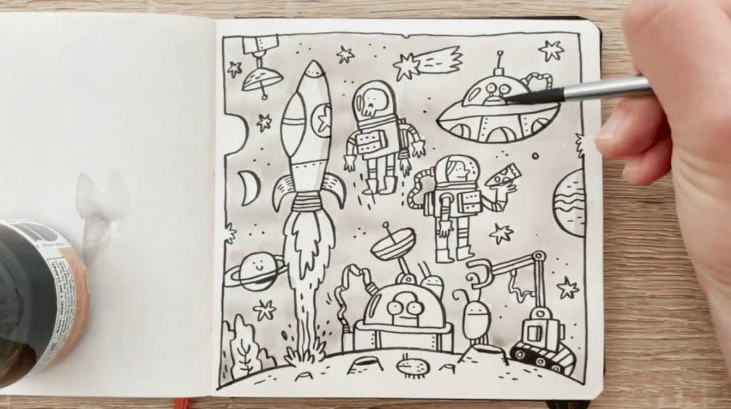 A hand holding a paint brush adds grey watercolor paint to a black and white drawing in a notebook. The drawing features multiple flying objects including a rocket, a flying saucer, astronauts, a comet and more.