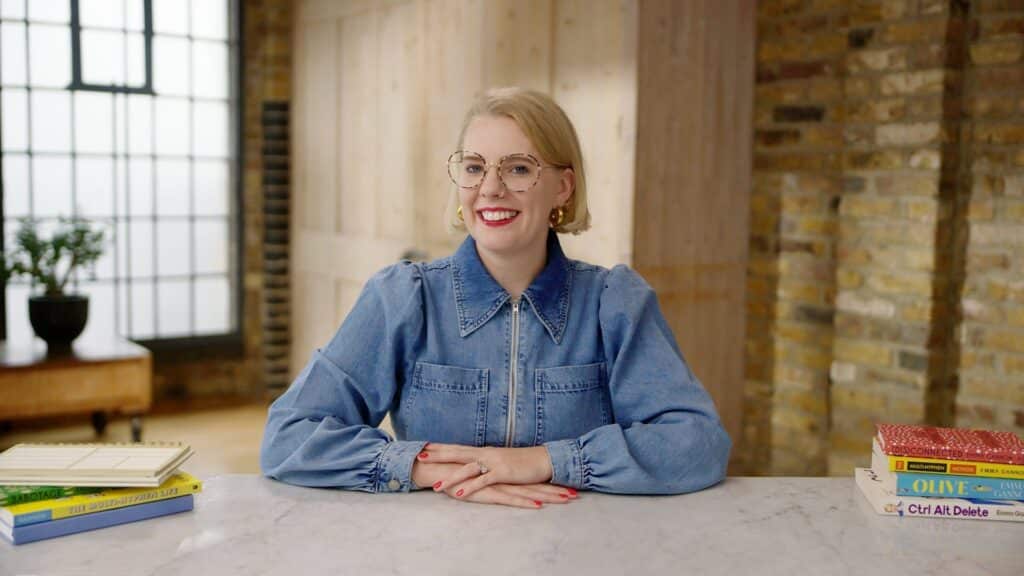 A blonde woman sits behind a desk and smiles at the camera. She is in an airy, bright studio.