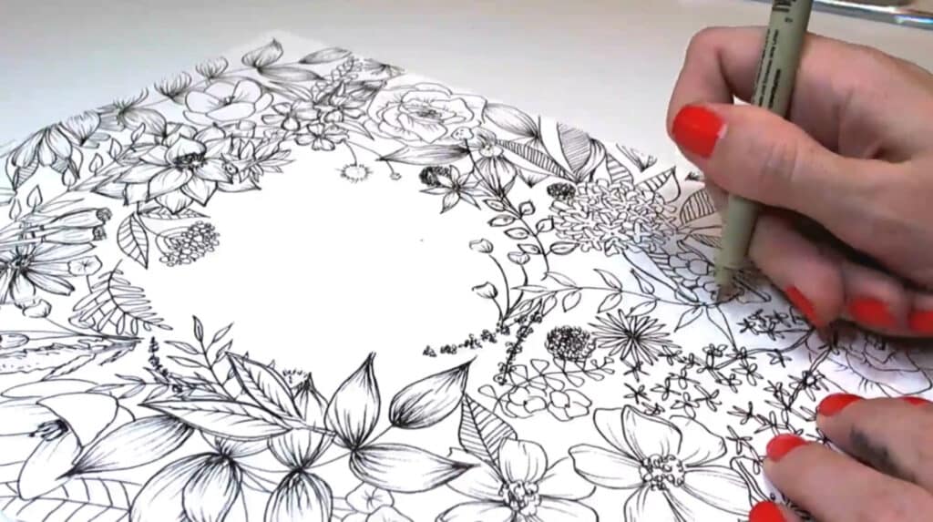 A woman’s hands using a Micron pen to complete a floral illustration on a white piece of paper. The flowers, vines and leaves cover nearly the entire page, except for a blank circular space in the middle.